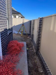 Construction site area with a tripod level set up beside a brick house wall.